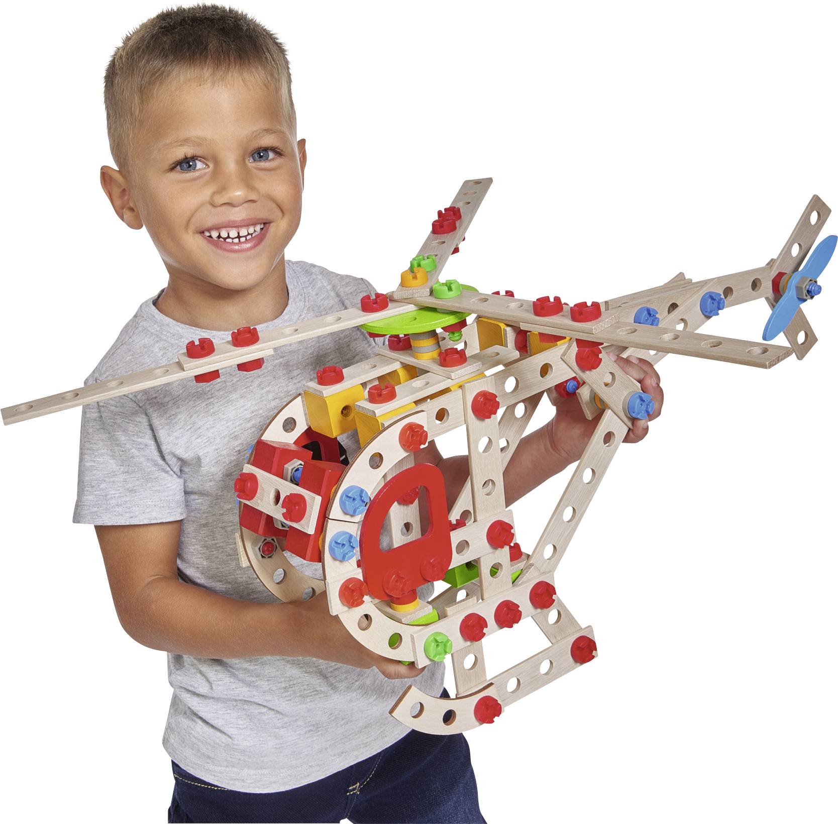 A child is smiling while holding a colourful wooden helicopter toy in their hands, against a white background.