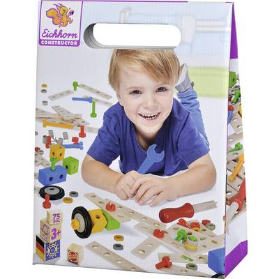 A child is lying on the floor, laughing and playing with a wooden building block set. In the background, colourful components and tools can be seen.