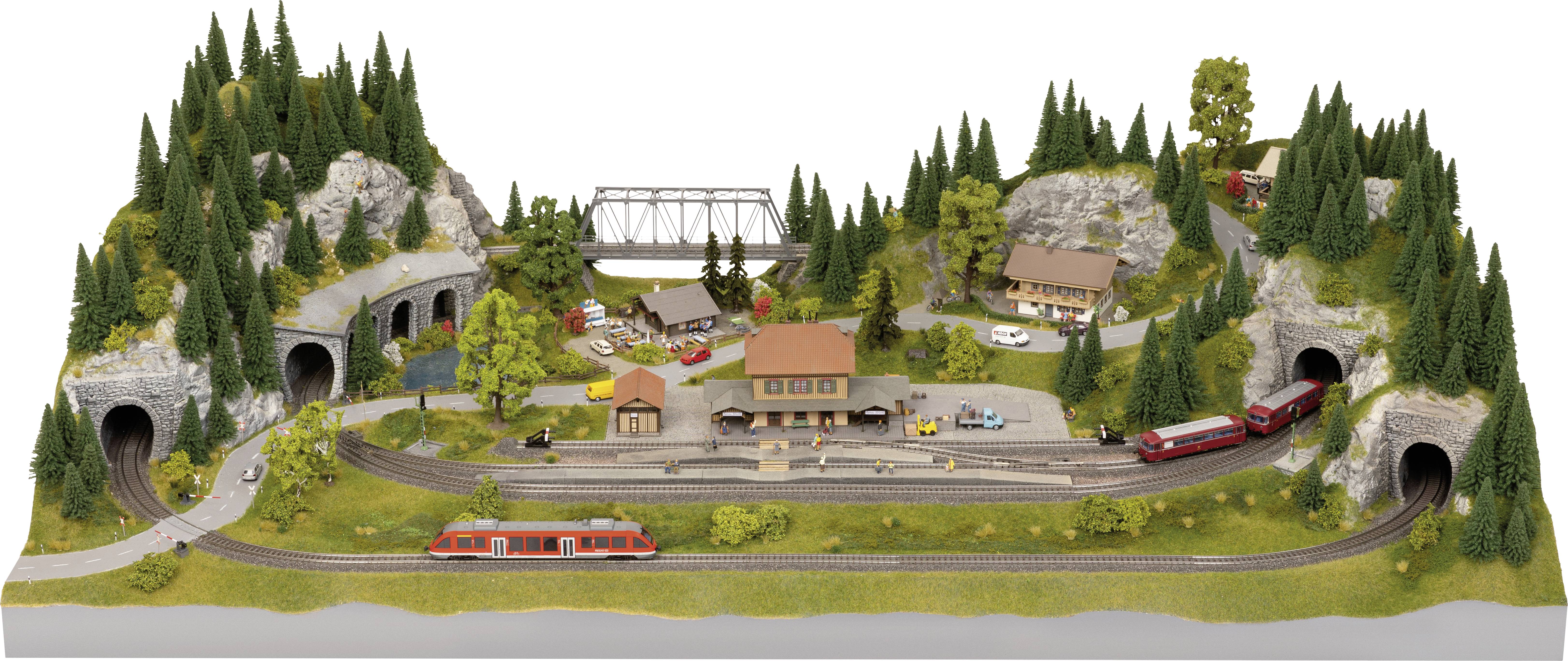 Model railway layout with mountainous landscape, multiple tunnel entrances, a station in the middle, and trains on tracks. Trees and buildings scattered throughout.