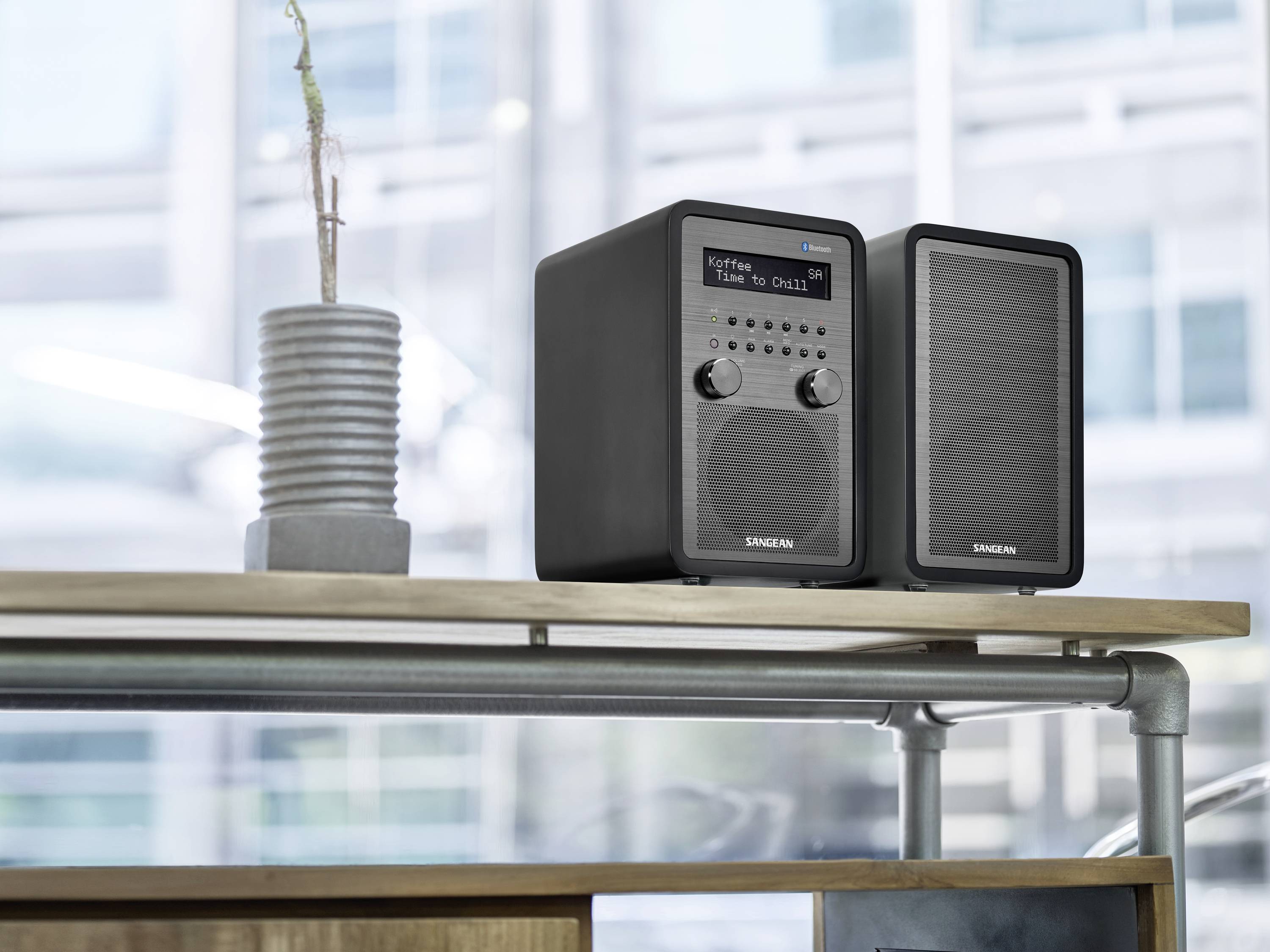 Compact black stereo system with display and speakers on a table, with a window and blurred exterior view in the background.