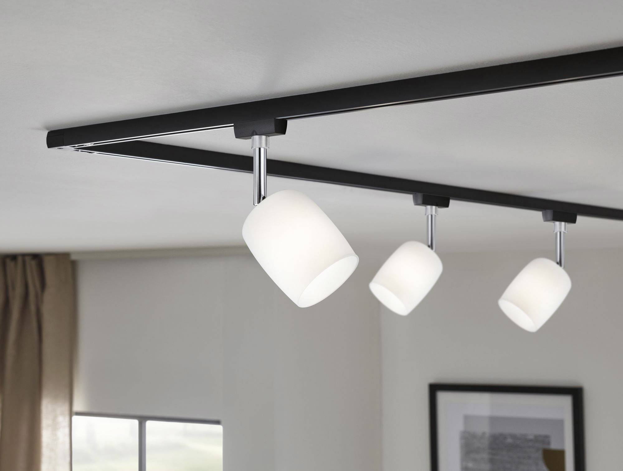 Ceiling light with three white, cylindrical lampshades mounted on a black track, installed on a light-coloured ceiling.