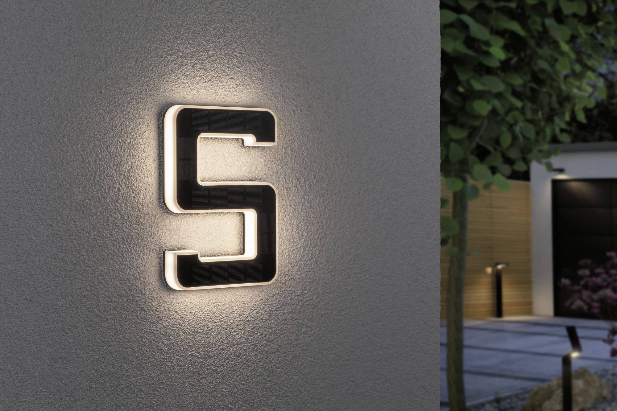 A backlit letter 'S' on a wall in evening light. Blurred trees and a modern courtyard are visible in the background.