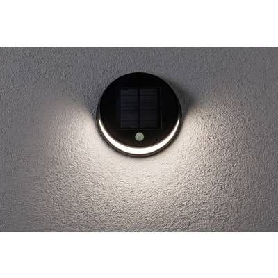 A round solar lamp mounted on a grey wall illuminates, with light radiating upwards and downwards.