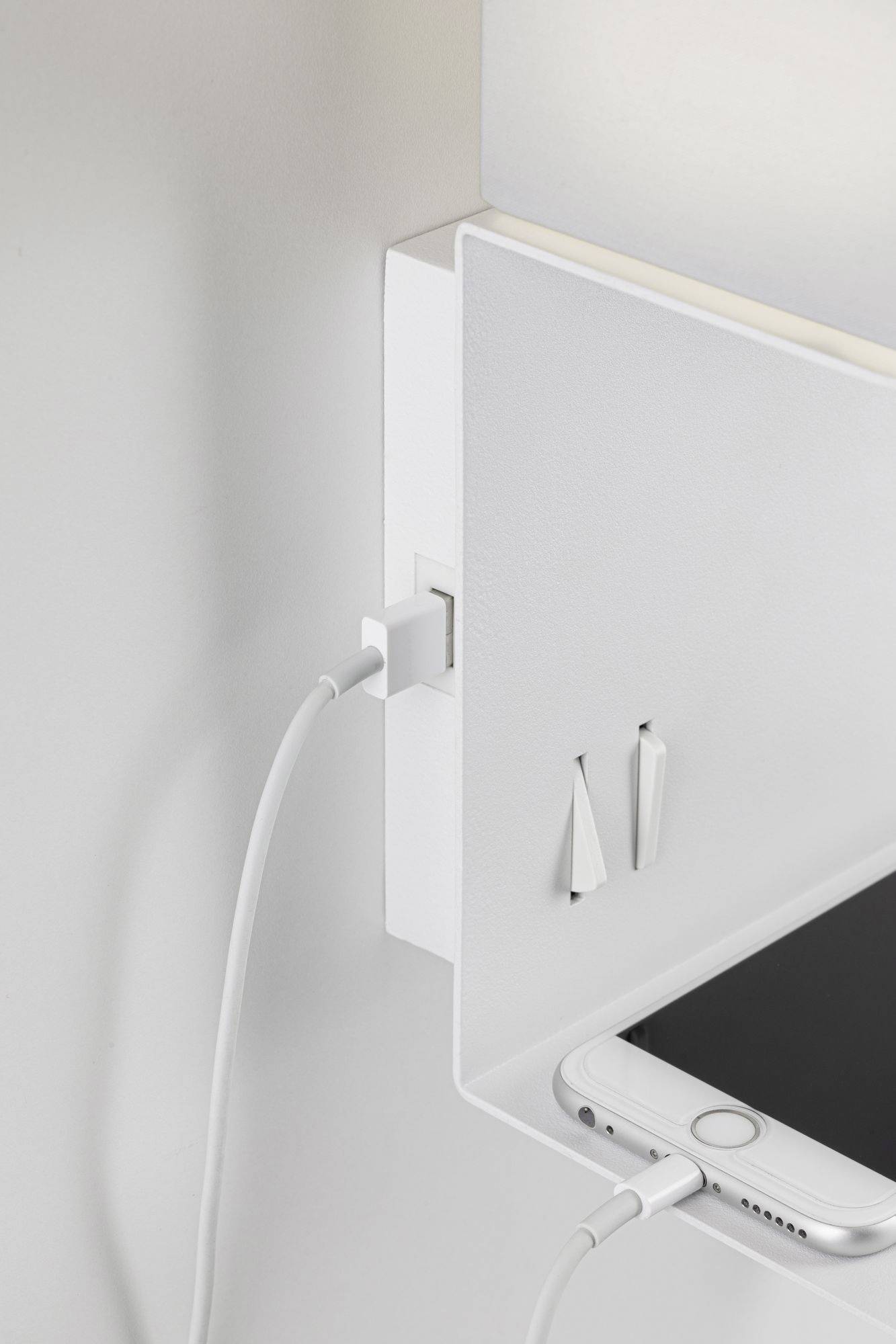 A smartphone is being charged at the bottom right of a white shelf. Another cable is plugged in at the top left. Two small decorations are hanging on the wall.