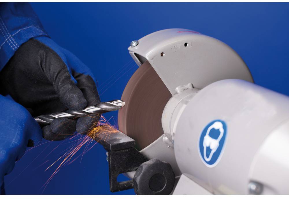 A person is grinding a drill bit on a grinding machine, with sparks flying. Protective clothing is visible, including blue gloves.