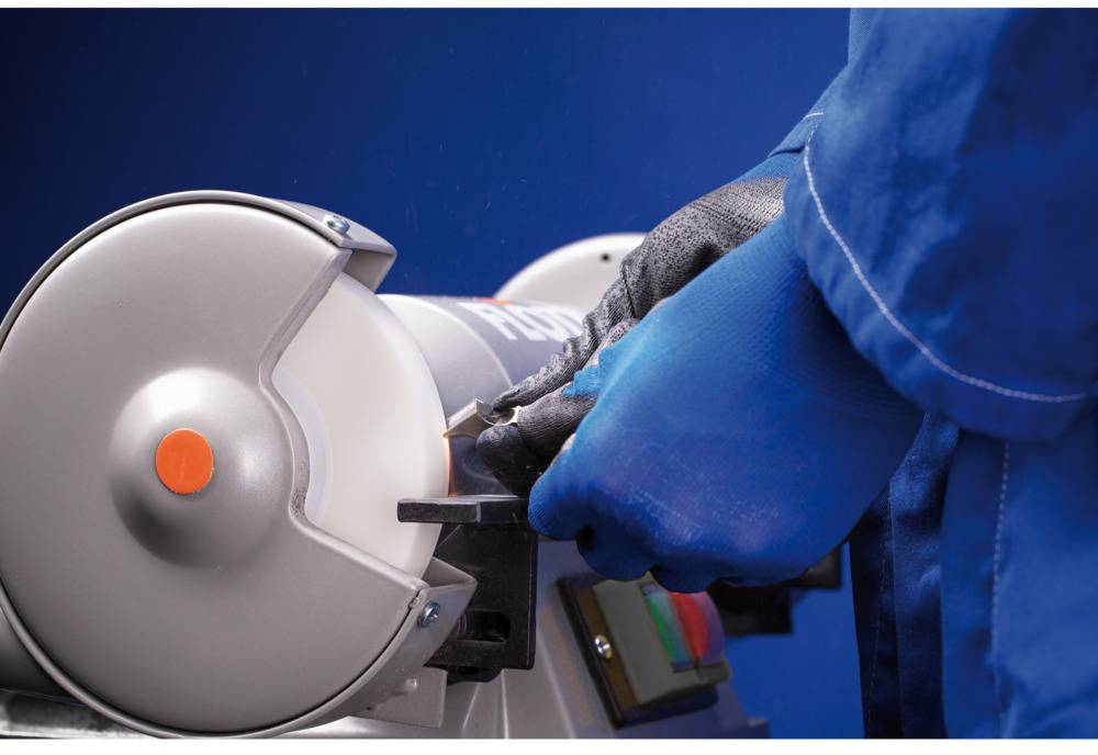 A person in blue protective equipment is grinding a metal piece on a grinding machine. The focus is on safety and precision.