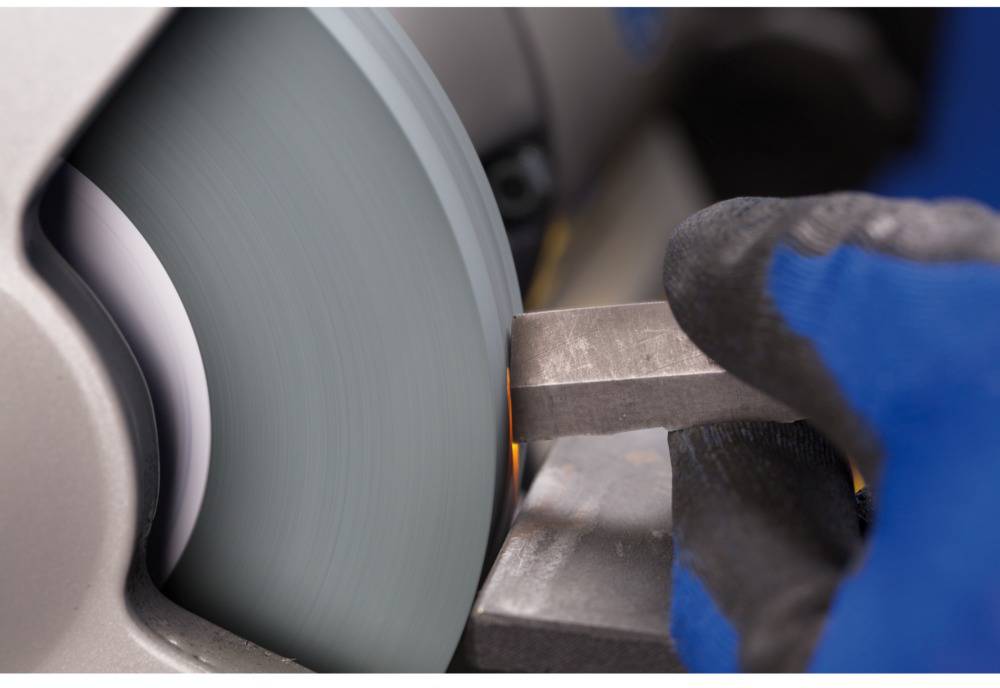 A hand wearing a glove grinds a piece of metal on a rotating grinding wheel. Sparks are generated during the grinding process.