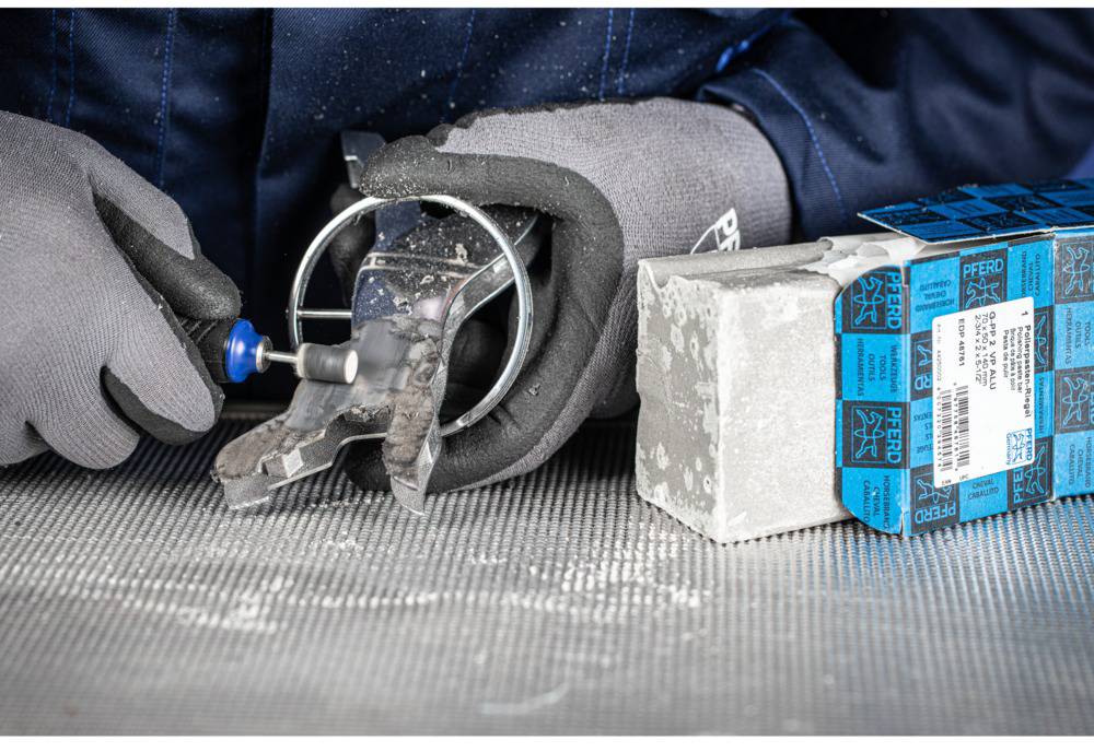 'Close-up of a person wearing protective gloves processing metal with a rotary tool. A stack of boxes is positioned to the right.'