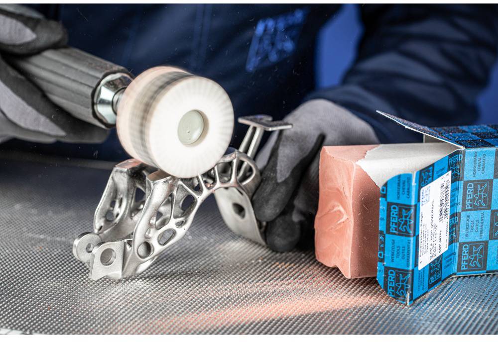 A person is polishing a complex-shaped metal part with a rotating brush. Next to them lies an opened package of abrasive materials.