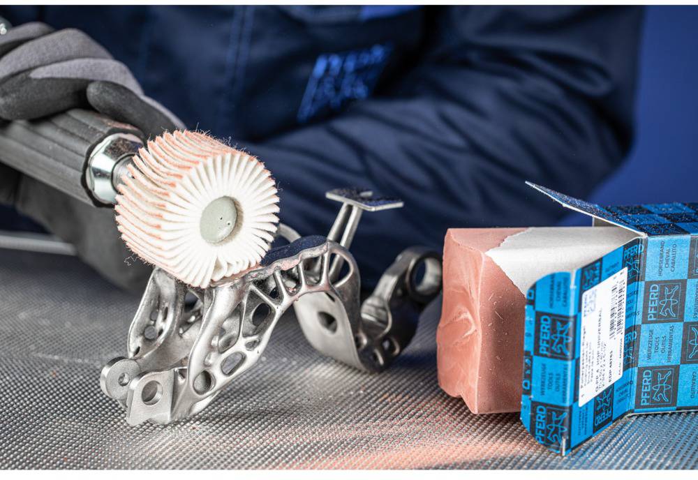 A person is polishing a metallic, intricate part with a rotating brush. A blue grinding block lies nearby on a table.