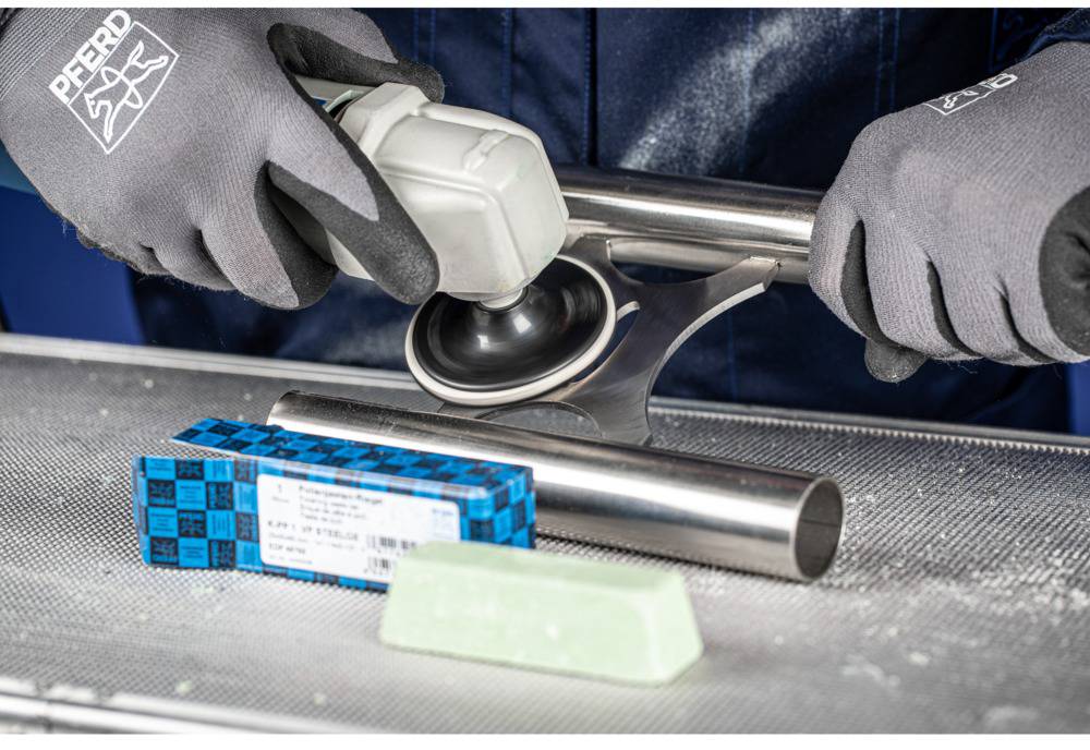 A person is polishing a metal rod with a machine. In the foreground, there is a pack of abrasive materials and a sanding block.