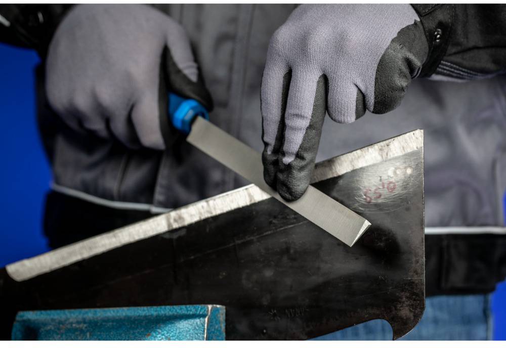 A person is wearing grey gloves and filing a metal edge with a file. The background is blue.