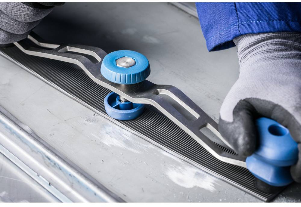 A person wearing grey gloves is using a blue-silver tool to straighten or shape a metal surface.