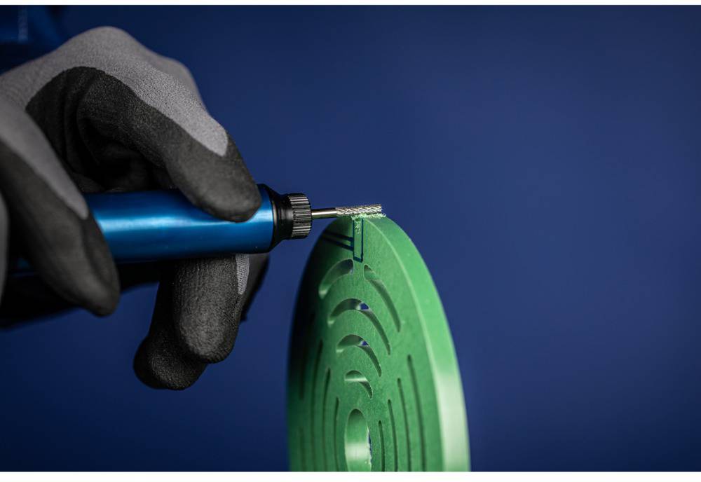 A person wearing a grey glove is holding a blue tool, working on a green, round object. The background is blue.
