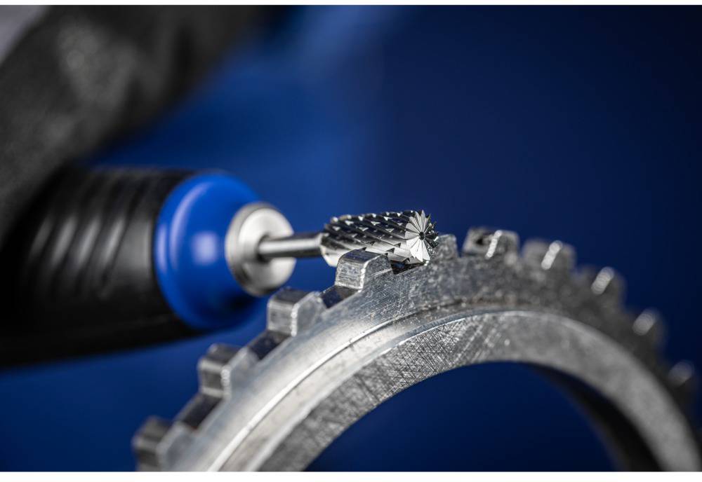 Close-up of a rotating milling drill bit machining the edge of a metal gear wheel. The background is blue, and a work glove is holding the tool.
