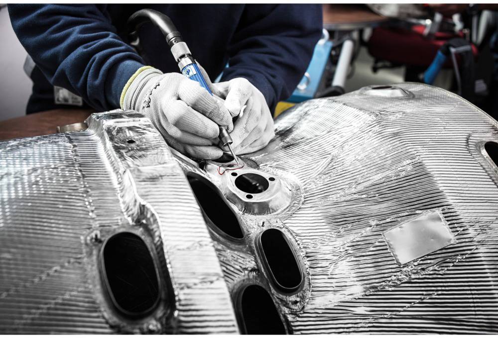 A person is working with safety glasses and gloves on a metallic component, holding a welding gun in their hand.