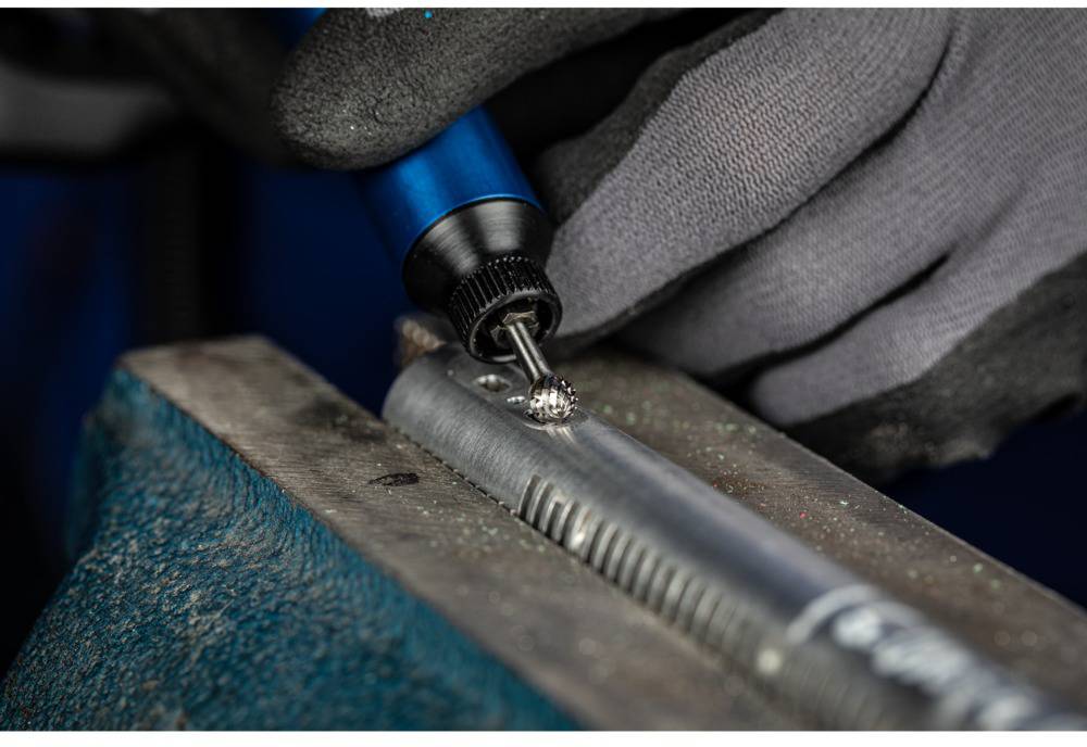 A person is welding metal on a vice with a blue soldering iron. Hands are wearing grey protective gloves.