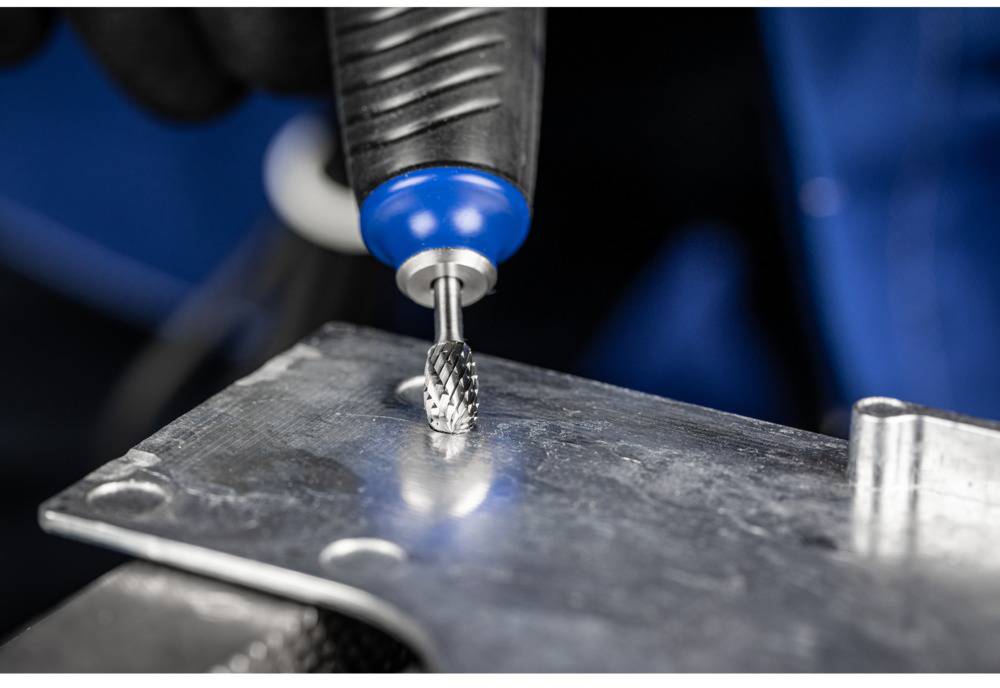 A hand holds a rotating grinding drill smoothing metal. The background is blurred and blue.