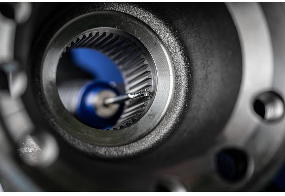 Close-up of a metal gear wheel with visible internal screws and teeth in focus. Blurred blue elements in the background.