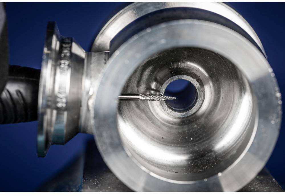 A view into a metal tube through which a drill bit precisely drills. Close-up, focus on the drill bit in the centre of the tube, background blurred.
