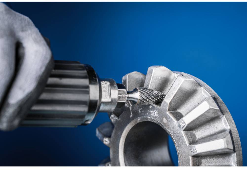 A hand holds a rotating tool working on a metallic gear. The background is blue.