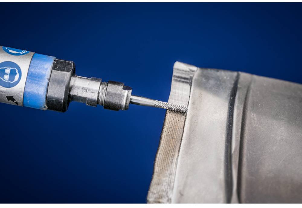 A metal tool smooths the edge of a metal piece against a blue background. The tool displays safety logos.