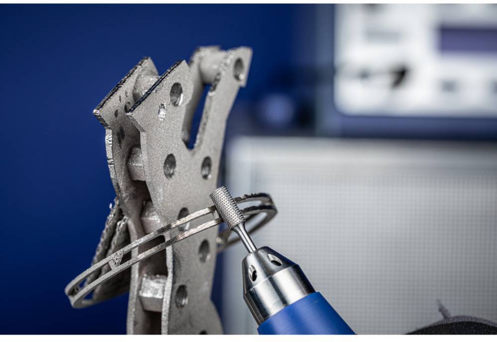 A metallic component is being machined with a rotating tool. A blue handle is visible. Background is out of focus.