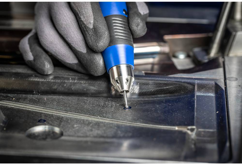 A hand wearing a glove holds a blue screwdriver. A screw is being screwed into a metal plate. The workplace looks industrial.