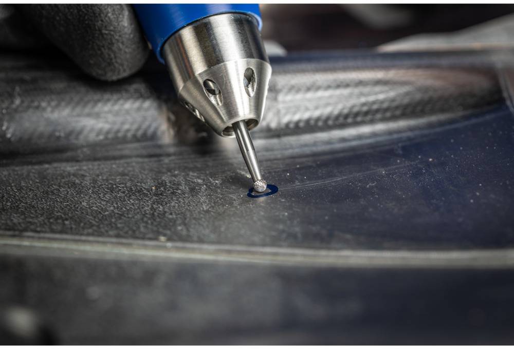 Close-up of a tool inserting a small metal rivet into a surface. The hand holds the tool precisely.