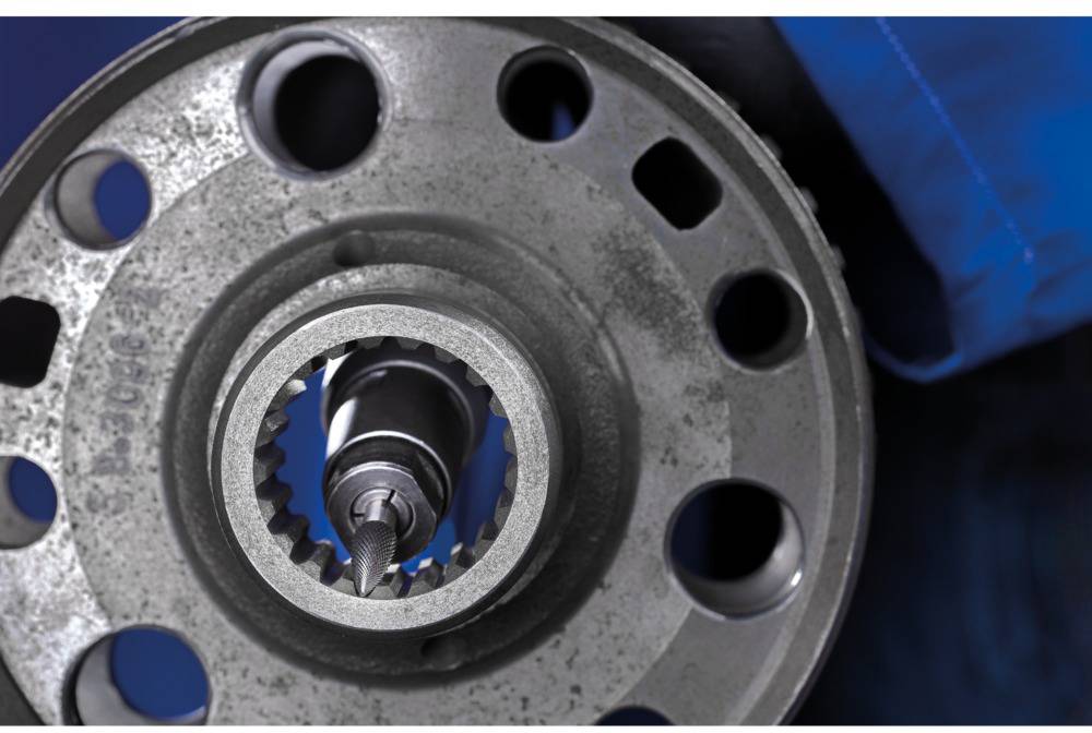 Close-up of a metal gear wheel with a drill head in the centre, performing a precise machining operation. In the background, a blue glove is visible.