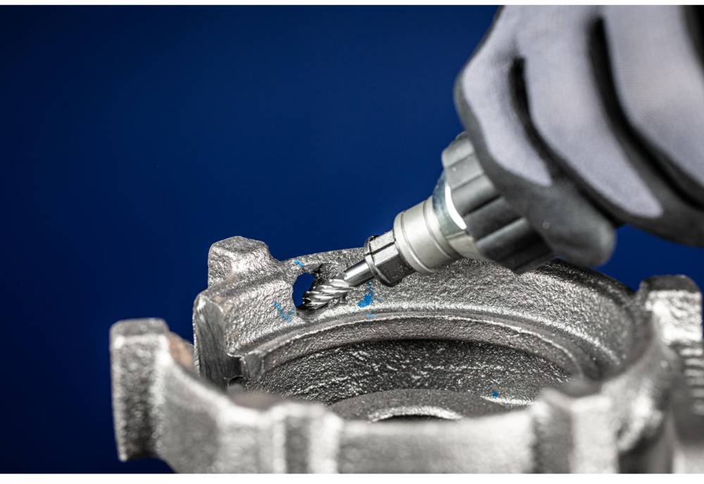 A gloved hand holds a rotating tool grinding metal from a component. Blue background.