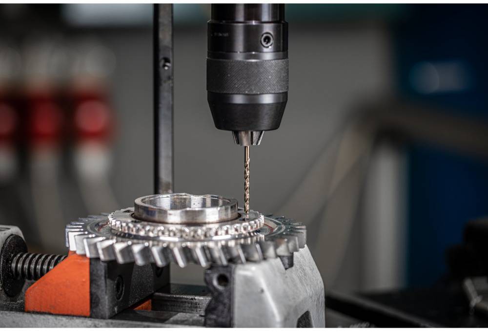 'Close-up of a drill bit boring into a metal cogwheel clamped in a vice. The background is blurred, with focus on the drilling work.'