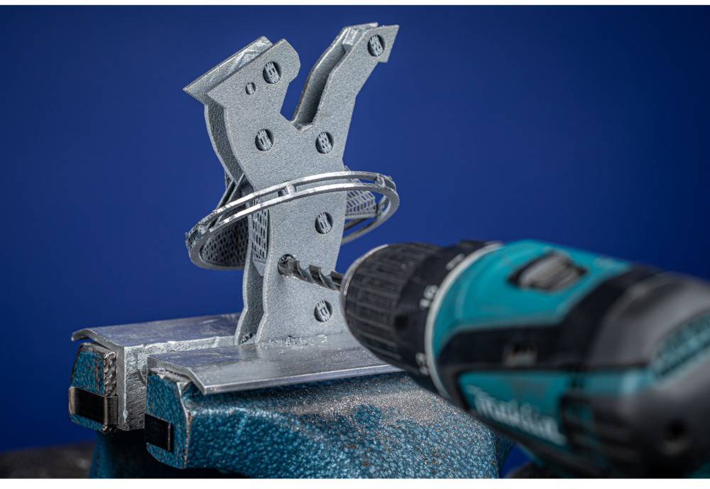 A drill is being used to make a hole in a grey metal piece shaped like the letter 'Y', which is secured in a mounting bracket.