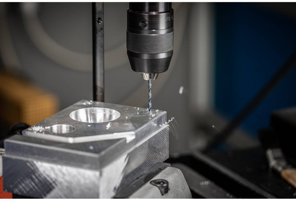 A drill is machining a rectangular metal block piece with circular recesses, surrounded by metal shavings.