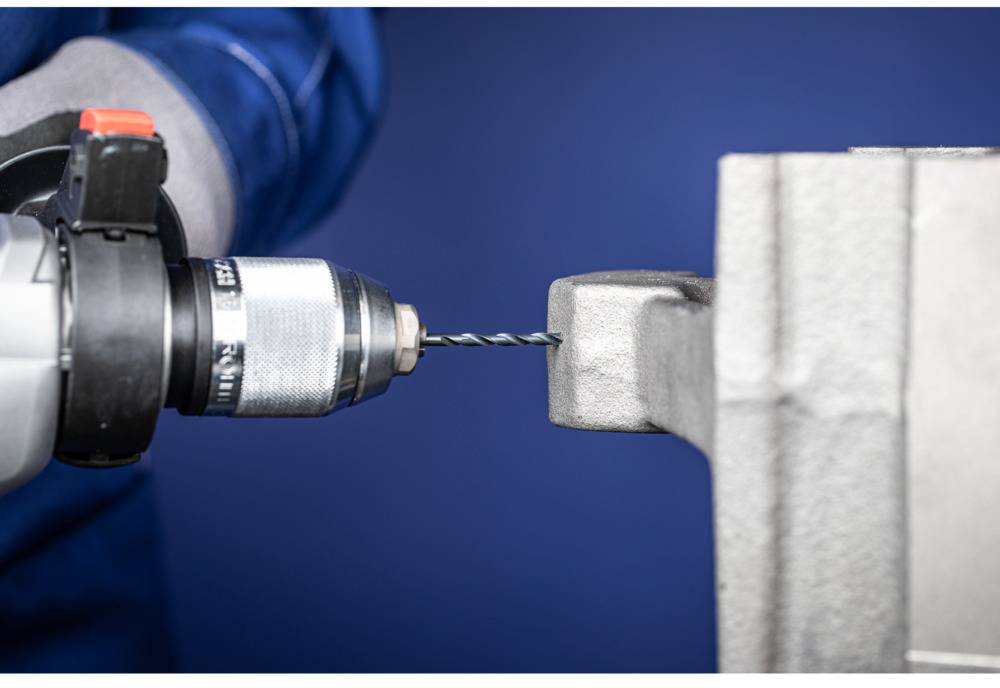 A drill is boring a hole into a grey piece of metal. The person is wearing blue workwear and gloves.
