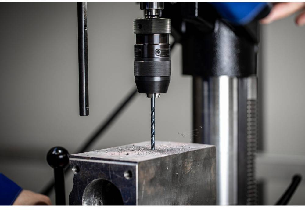 A drill is boring a hole into a metal block. The drill bit is rotating and metal shavings are visible.