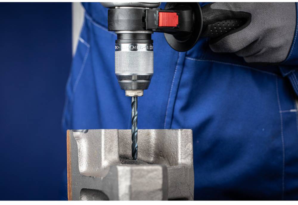Close-up of a person wearing blue workwear using an electric drill to bore into a metal block.