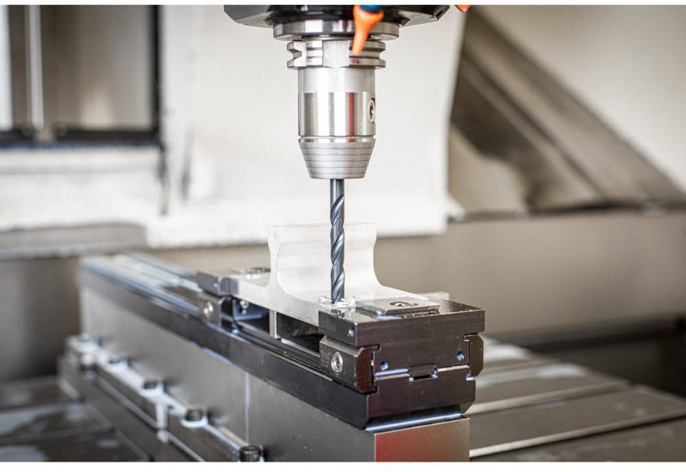A drill bit is machining a piece of metal on a CNC machine, clearly visible in the foreground, with focus on the drill tip.