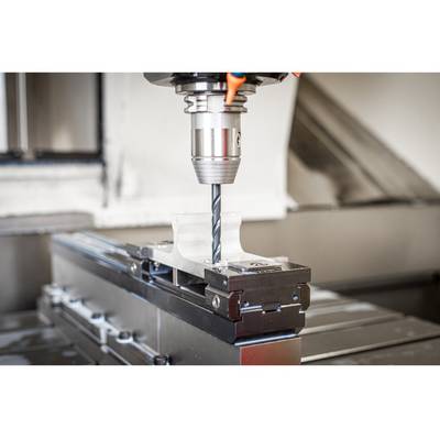 A drill bit is machining a piece of metal on a CNC machine, clearly visible in the foreground, with focus on the drill tip.