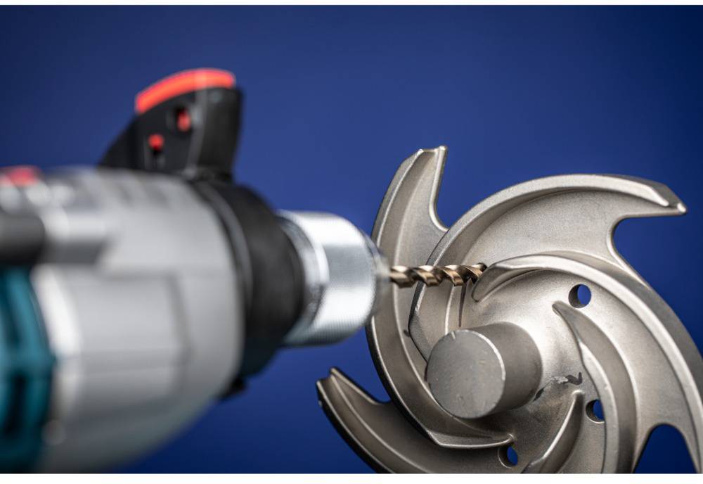 Close-up of a drill bit drilling into a metallic machine component. The part has curved blades and a polished surface.