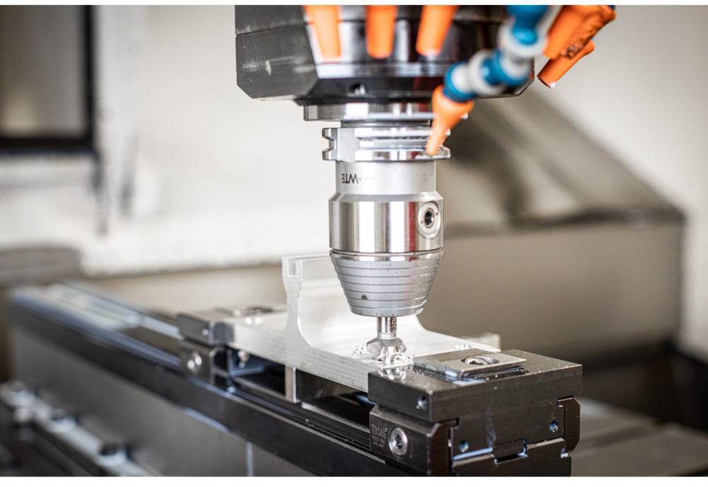 A CNC milling machine is machining a metal workpiece. The milling head is positioned close to the material and is processing it with precision. Several coolant hoses are visible.