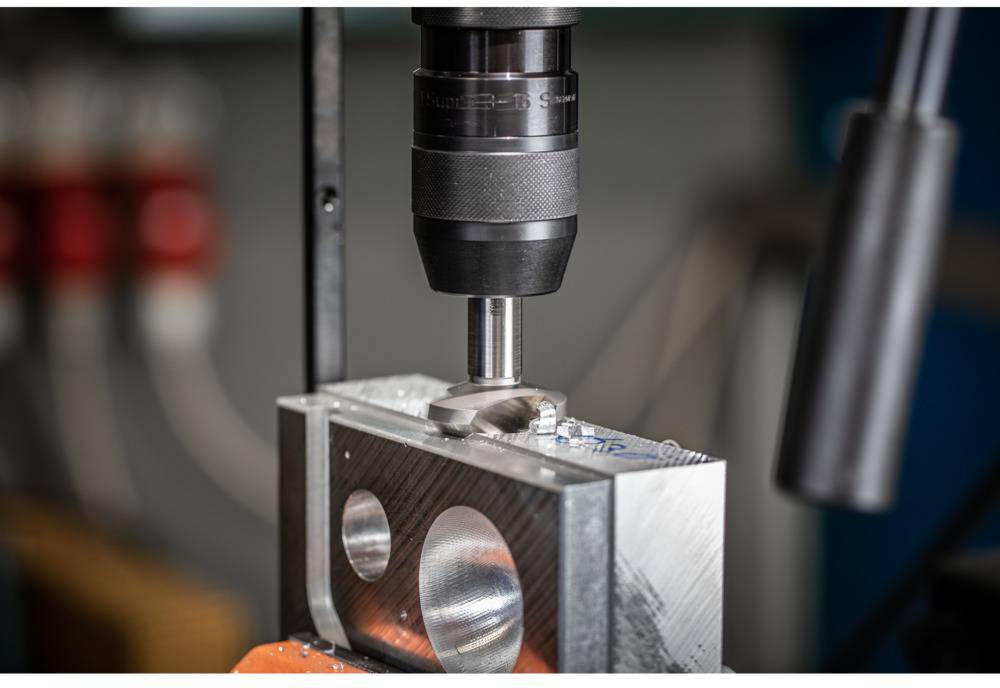 Drill machine processing a metal block with circular recesses. Focus on the drill bit and workpiece, with an industrial environment in the background.