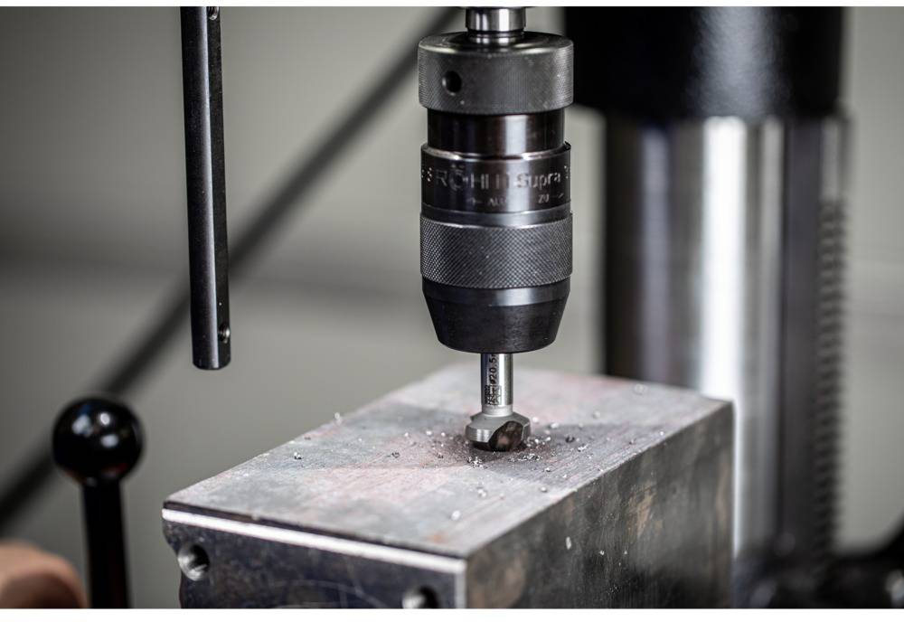 Close-up of a drill boring a hole into a piece of metal. Metal shavings are visible on the surface.
