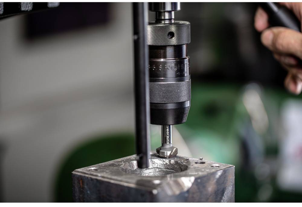 A metal block is being machined with a drill. A drill bit descends into the metal while a person holds the machine.