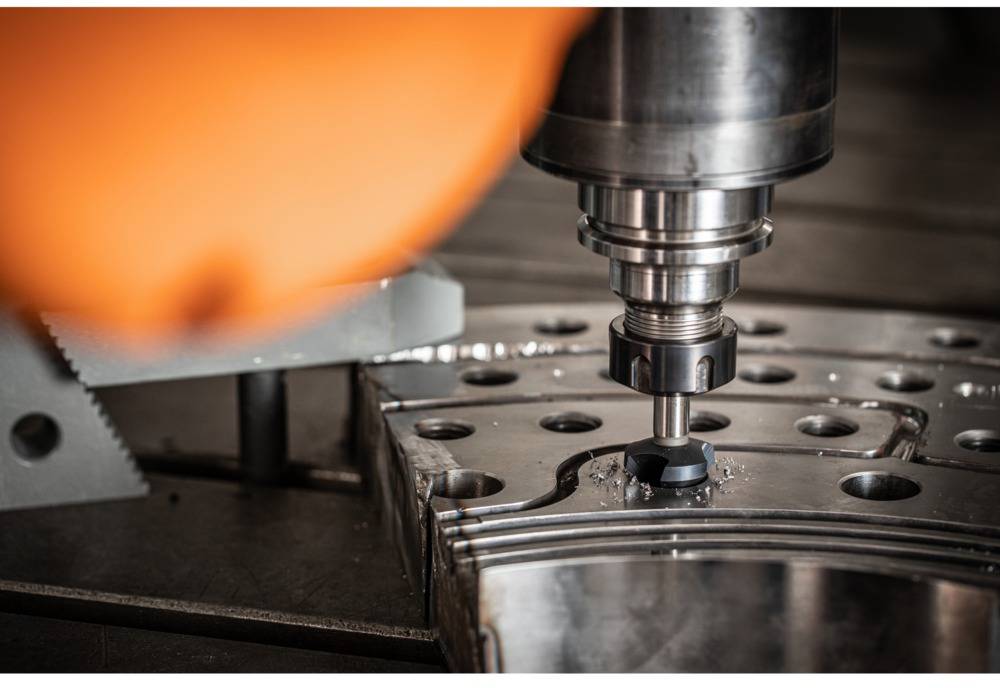 A CNC machine precisely machines a metal piece with multiple holes. In the foreground, a red-orange object is visible.
