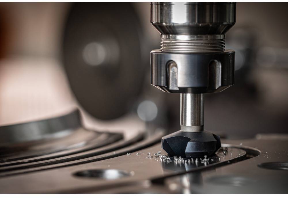 A milling machine is machining a metal surface, creating swarf. In the background, blurred machine components are visible.