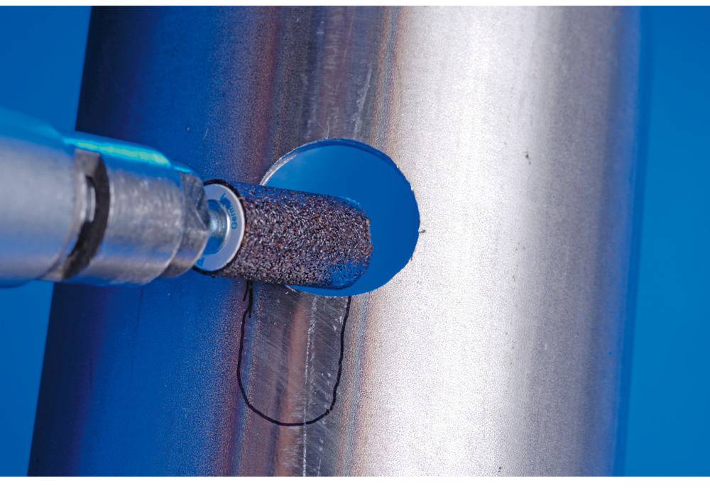 A metal pipe is being smoothed with a rotating grinding attachment, which is polishing a round opening. Blue background.