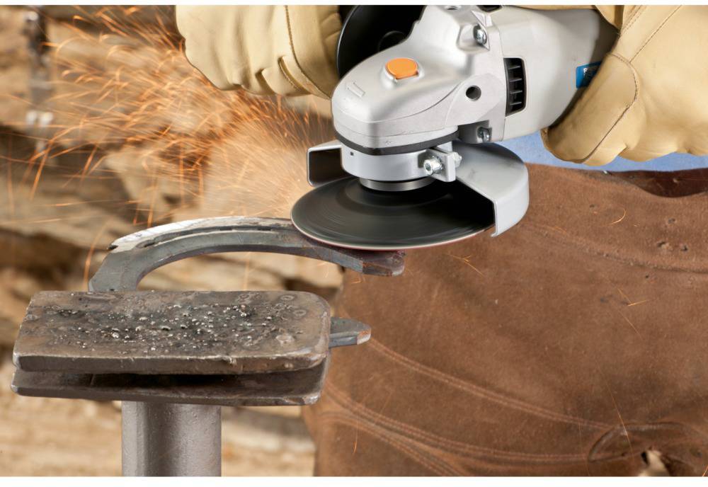 A person is grinding metal with an angle grinder, sparks flying. Protective clothing and clamp are visible for safe handling.