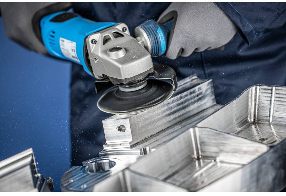A person wearing gloves is using a grinder to work on metal. Metallic shavings are flying, with detailed work being carried out on a machine.
