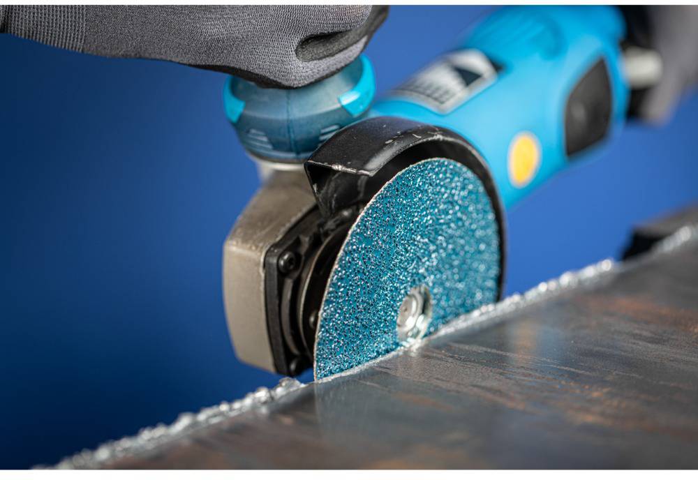 A blue electric tool with a grinding disc is sanding a metal edge. Sparks and a gleaming surface emphasise the machining process.